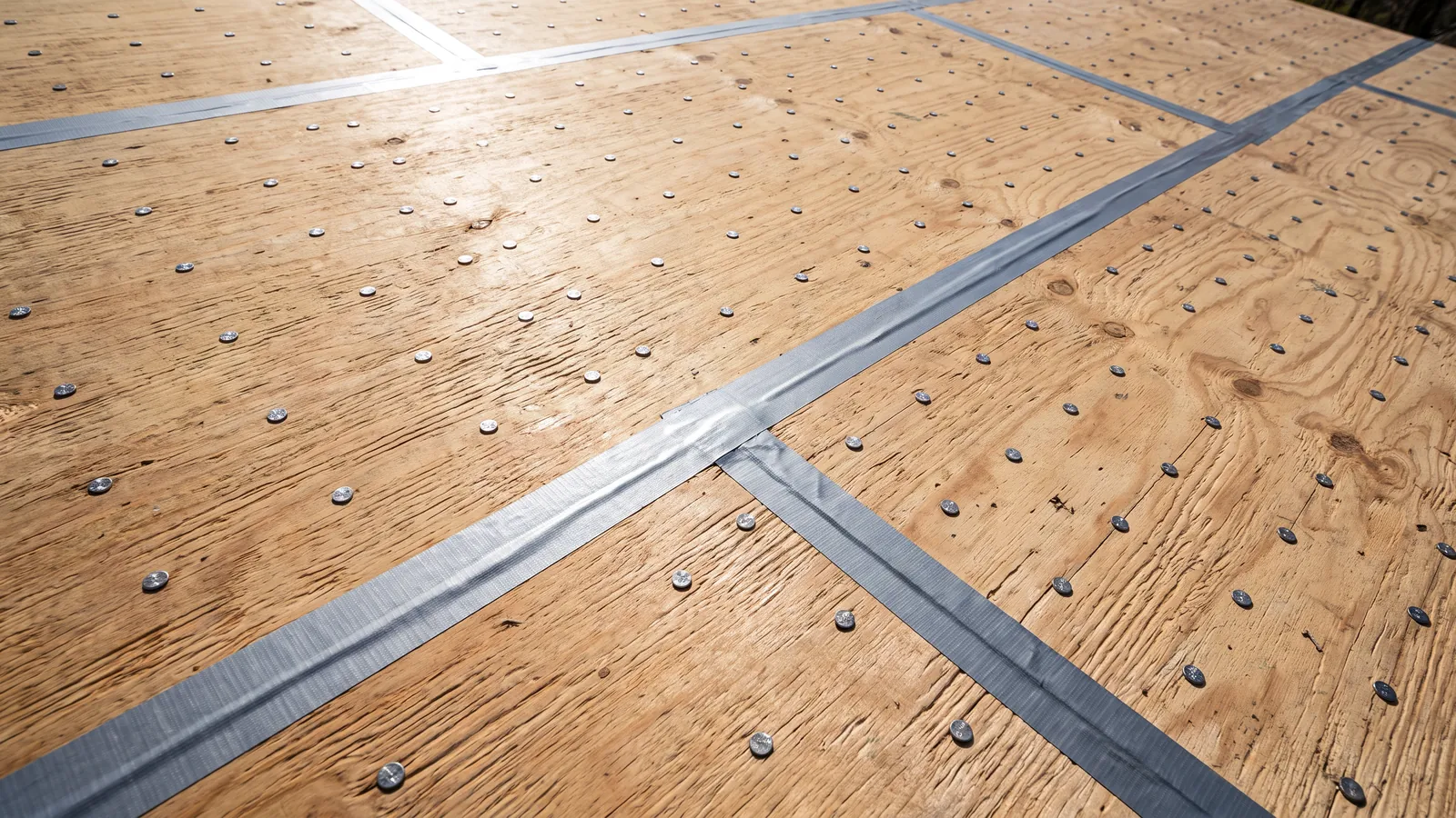 Bare roof decking with every seam sealed by tape before underlayment, ring-shank nails driven in a tight pattern across the plywood